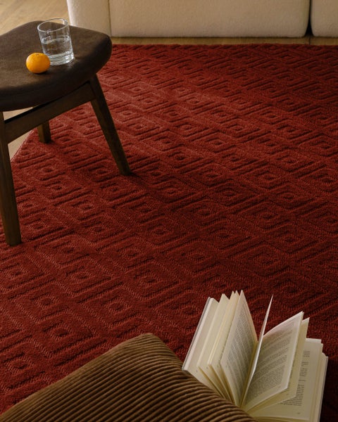 Strukturierter roter Teppich mit geometrischem Quadratmuster in einem hellen Wohnzimmer mit Hocker, Buch und Kissen.