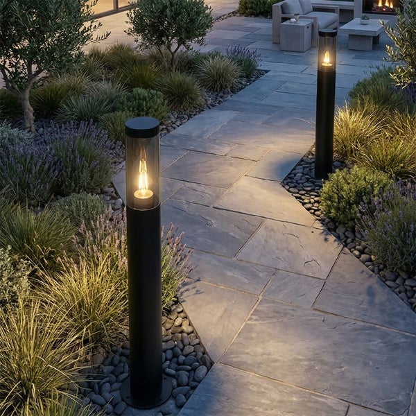 Zwei schwarze Außenstandleuchten mit warmem LED Licht auf einem Gartenweg aus grauen Steinplatten gesäumt von Pflanzen und Kieselsteinen.