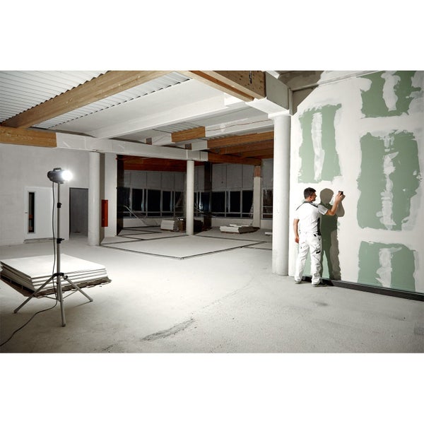 Handwerker beim Verspachteln einer Trockenbauwand in einem hellen Raum mit Baustrahler, Gipsplatten und sichtbaren Deckenbalken.