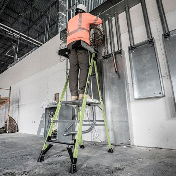 Ein Arbeiter auf einer grünen Plattformleiter installiert Kabel an einer Wand in einer hellen Industriehalle auf einer Baustelle.