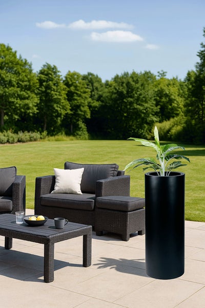 Hoher, zylindrischer Pflanzkübel in Schwarz auf einer Terrasse mit Rattan-Gartenmöbeln und Blick auf eine große Rasenfläche.