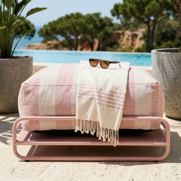 Gestreifter Outdoor-Pouf auf rosa Metallgestell mit Handtuch und Sonnenbrille, auf einer Terrasse an einem Pool.