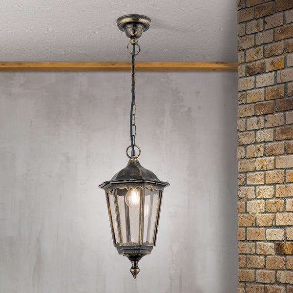 Antike Hängeleuchte in Laternenform aus Metall und Glas mit gold-schwarzer Patina an einer Kette vor einer Ziegelwand.