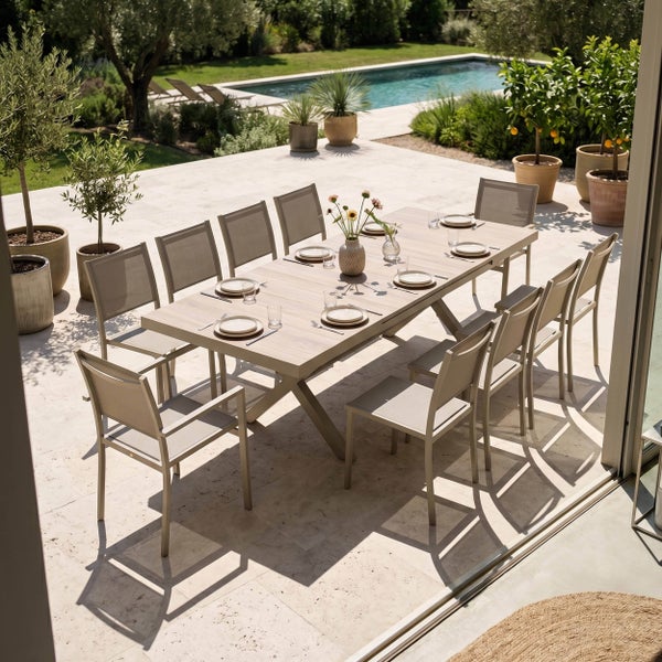 Gedeckter Gartentisch aus Holz mit Keramiktellern und Besteck auf einer hellen Terrasse mit Pool im Hintergrund.