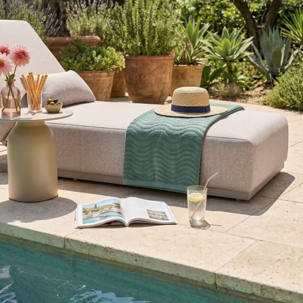 Helle Szene mit gepolsterter Gartenliege am Pool, Beistelltisch, Magazin, Handtuch, Strohhut und Pflanzen in Terrakottatöpfen auf einer Steinterrasse.