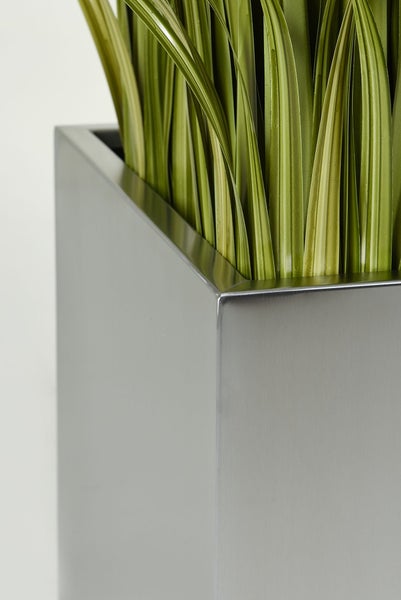 Detailaufnahme eines quadratischen silbernen Pflanzkübels mit künstlichen grünen Grashalmen.