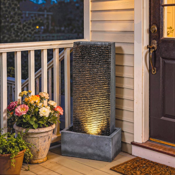 Moderner rechteckiger Gartenbrunnen mit strukturierter Wasserwand und LED-Beleuchtung, platziert auf einer Terrasse neben einem Hauseingang.
