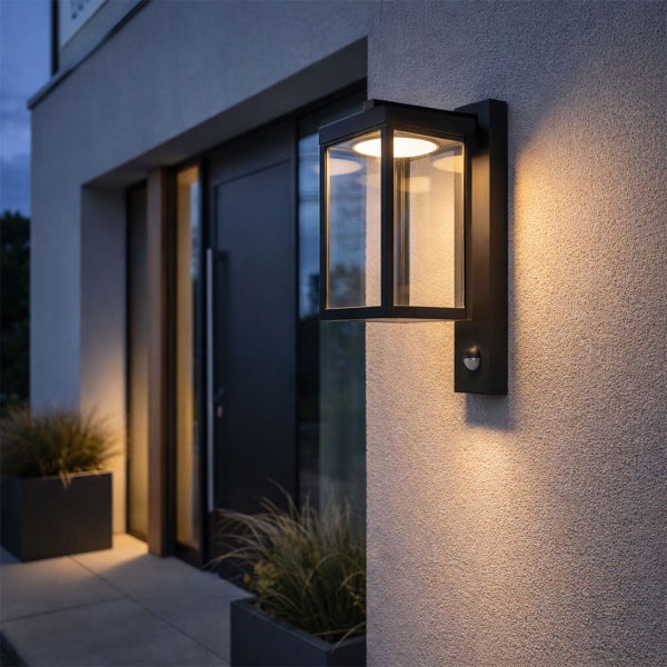 Moderne schwarze Außenwandleuchte mit Bewegungsmelder und Glaselementen, montiert neben einem Eingangsbereich, strahlt warmes Licht aus.