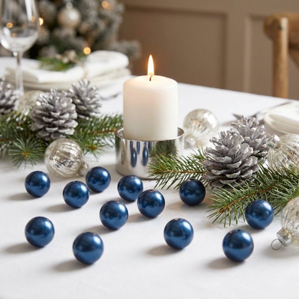 Weihnachtliche Tischdekoration mit blauen Dekokugeln, silbernen Tannenzapfen und einer brennenden Stumpenkerze auf einer weißen Tischdecke.