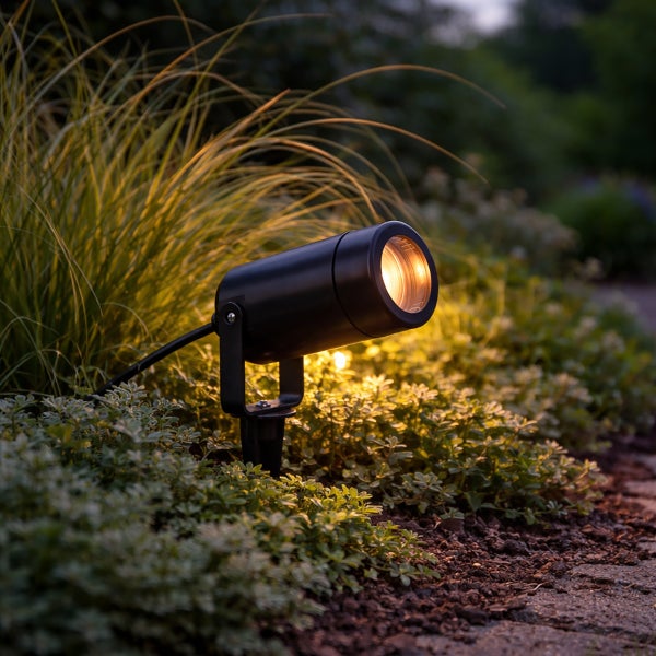Schwarzer Gartenstrahler mit Erdspieß beleuchtet Pflanzen im Garten mit warmem Licht.