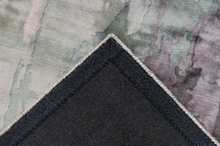 Detailansicht eines Teppichs mit abstraktem Muster in Grün, Grau und Violett, umgeklappte Ecke zeigt die dunkle Rückseite.