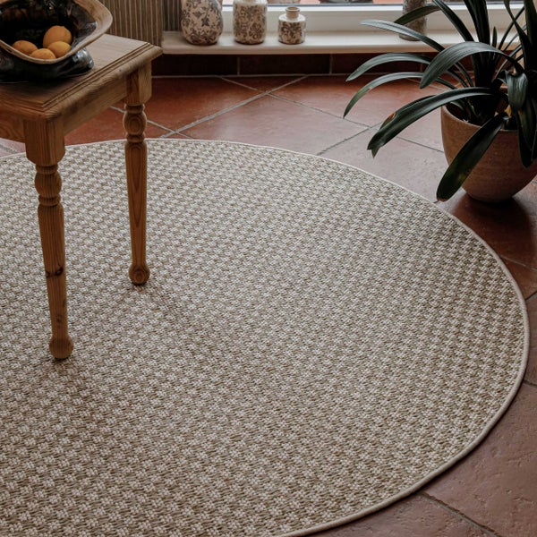 Runder gewebter Teppich in Sisal-Optik auf einem Fliesenboden neben einem Holzhocker und einer Zimmerpflanze.