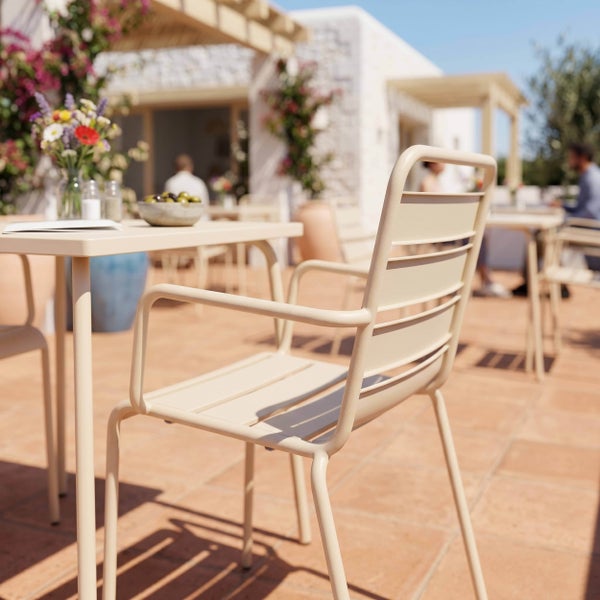 Moderner Gartenstuhl aus Metall mit Armlehnen an einem Tisch auf einer hellen Terrasse mit Fliesenboden und Steinmauer.