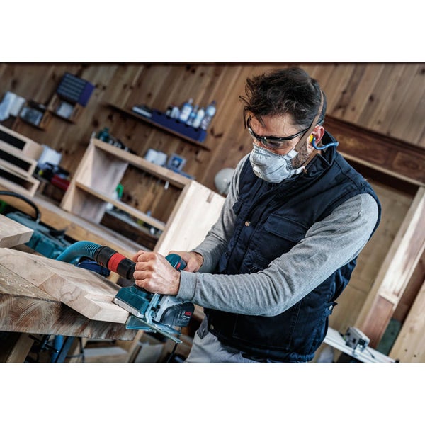 Ein Mann hobelt ein Stück Holz mit einem Elektrohobel und trägt dabei eine Maske, eine Schutzbrille und Ohrstöpsel.