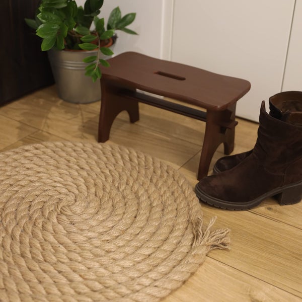 Ein runder Teppich aus gewundenem Naturseil liegt auf einem Holzboden neben einem dunkelbraunen Tritthocker aus Holz und braunen Stiefeln.