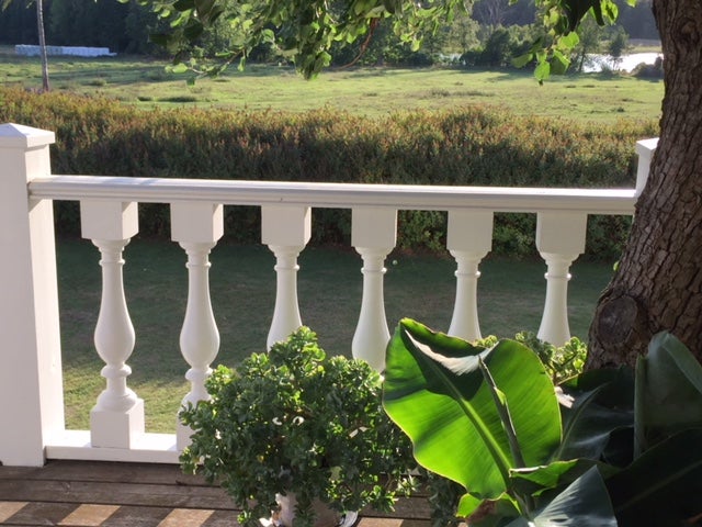 Weißes Balkongeländer im klassischen Balustraden-Stil auf einer Holzterrasse mit Blick ins Grüne und Topfpflanzen im Vordergrund.