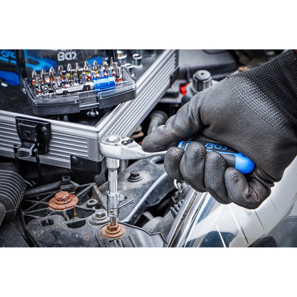 Eine Hand mit Handschuh benutzt eine Knarre mit Steckschlüsselsatz an einem Motor, ein Bit-Set liegt daneben.