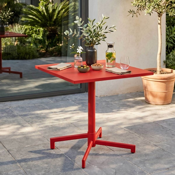 Roter quadratischer Bistro-Tisch auf einer hellen Steinterrasse mit Olivenbaum im Terracotta-Topf.