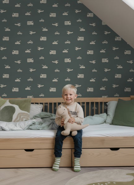 Dunkelgrüne Tapete mit Fahrzeugmuster wie Feuerwehrautos und Hubschraubern in einem Kinderzimmer hinter einem Holzbett.