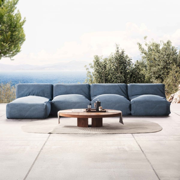 Modulares Outdoor-Sofa in Blau auf einer Terrasse mit Meerblick, runder Holztisch auf hellem Teppich.