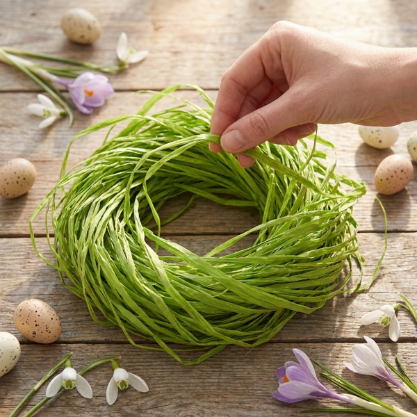Hand formt einen dekorativen Osterkranz aus Bast auf einem rustikalen Holztisch umgeben von Frühlingsblumen und gesprenkelten Ostereiern.
