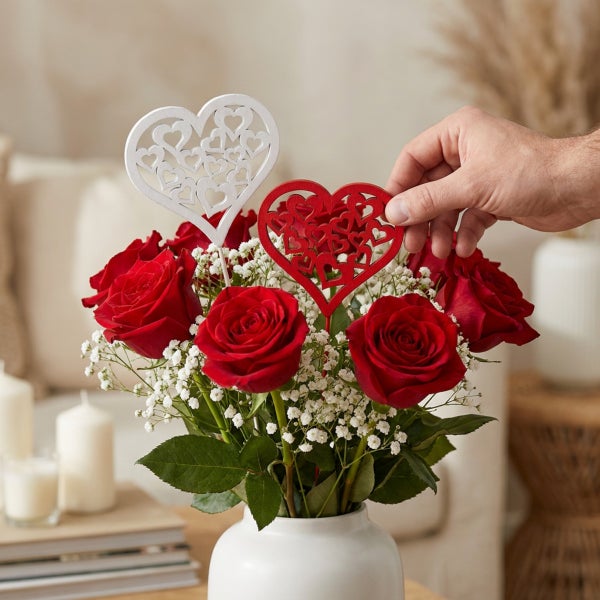 Ein Blumenstrauß aus Rosen und Schleierkraut in einer Vase, dekoriert mit zwei herzförmigen Steckern aus Holz.