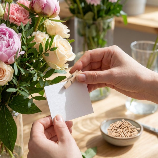 Hände halten eine leere Grußkarte mit einer Holzklammer neben einem Blumenstrauß aus Pfingstrosen und Rosen auf einem Holztisch.