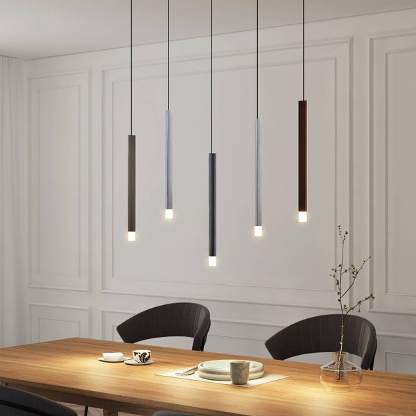 Fünf schlanke röhrenförmige Pendelleuchten in verschiedenen Metallfarben hängen in verschiedenen Höhen über einem Holztisch in einem hellen Raum.