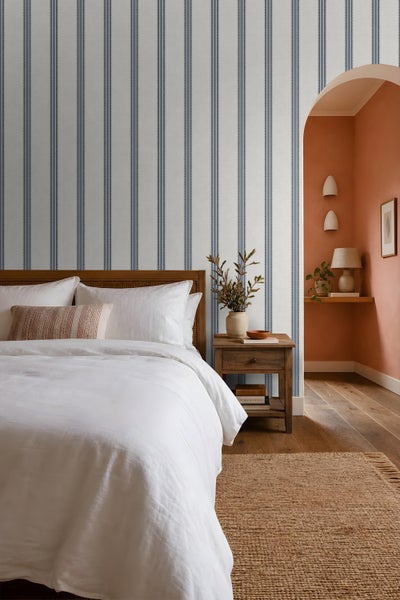 Schlafzimmer mit Vliestapete mit Längsstreifenmuster, Holzbett, Nachttisch und angrenzender Nische in warmen Tönen mit Tischleuchte.