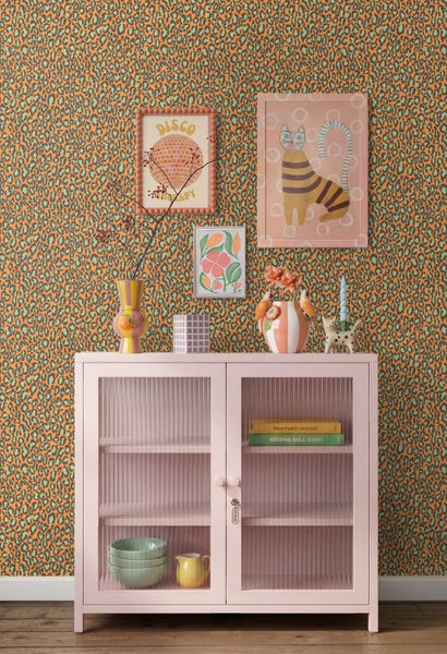 Tapete mit Leopardenmuster an der Wand hinter einer rosa Vitrine. Eklektische Raumgestaltung mit bunten Dekorationsartikeln und Bildern.