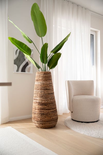 Große geflochtene Bodenvase mit grüner Pflanze in einem hellen Wohnzimmer neben einem Sessel.