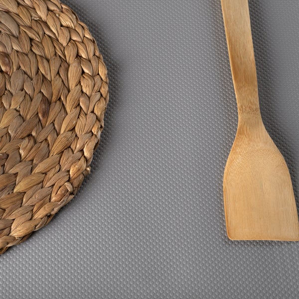 Ein Pfannenwender aus Holz und ein geflochtenes Tischset auf einer grau strukturierten Schubladeneinlage.