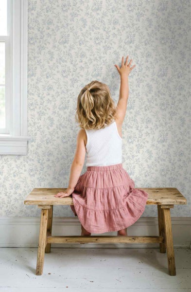 Helle Vliestapete mit filigranem Blumenmuster in einem Kinderzimmer, ein Kind sitzt auf einer rustikalen Holzbank davor.