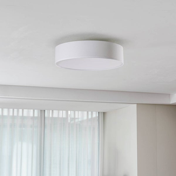 Runde Deckenleuchte mit Textilschirm an einer weißen Decke in einem hellen Raum mit Fenstervorhang.