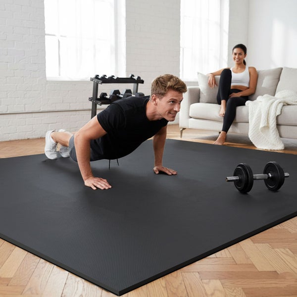 Mann macht Liegestütze auf einer Trainingsmatte mit einer Frau, die auf einem Sofa sitzt und einer Hantel auf dem Boden.