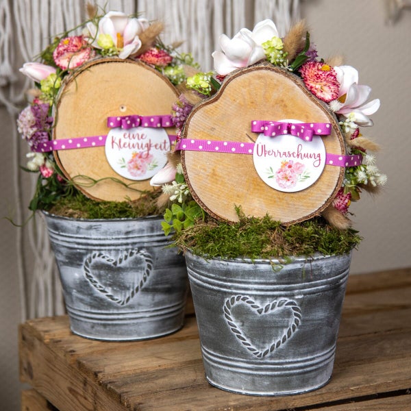 Zwei dekorative Holzscheiben mit Blumenschmuck und Geschenkanhänger in grauen Metalltöpfen mit Herzprägung auf einem Holztisch.
