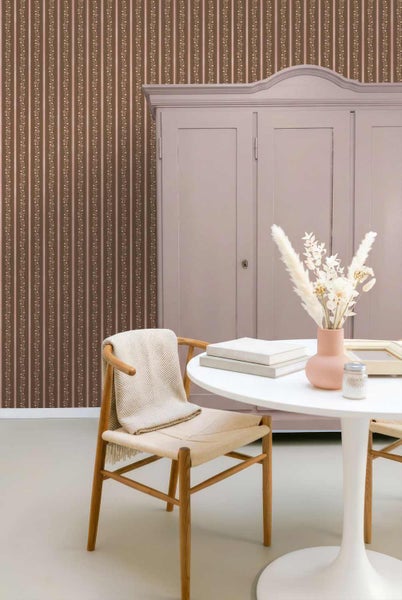Zimmer mit braun-beige gestreifter Blumentapete, Holzstuhl mit Decke, weißem Rundtisch mit Trockenblumen und einem altrosa Schrank.