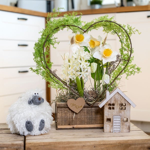 Dekorative Anordnung mit Herz, Narzissen, Hyazinthen, Holzhaus und Stoffschaf auf einem Holztisch