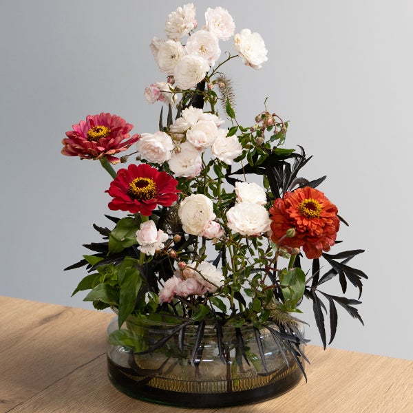 Blumenarrangement mit weißen Rosen, roten und rosa Zinnien sowie dunklem Blattwerk in einer flachen Glasschale auf einer Holzoberfläche.