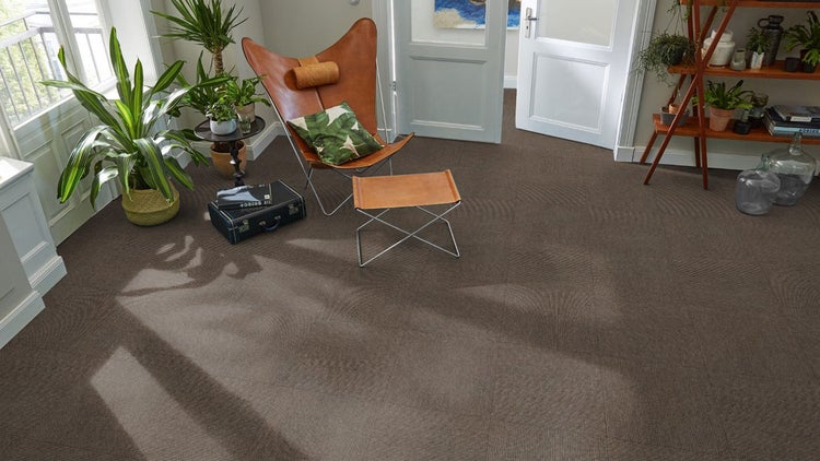 Braune Teppichfliesen in einem hellen Wohnzimmer mit Ledersessel, Pflanzen und Holzregal.