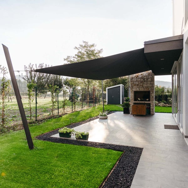 Modernes dunkles Sonnensegel über einer gefliesten Terrasse mit Gartenkamin und gepflegter Grünfläche.