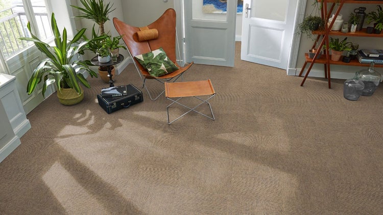 Heller Wohnraum mit strukturierten braunen Teppichfliesen, einem Butterfly-Sessel aus Leder, Zimmerpflanzen und einem Holzregal.