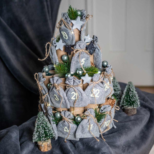 Dreistöckiger Adventskalender mit Stoffbeuteln und Zahlenaufdruck, dekoriert mit Kugeln, Tannenzweigen und kleinen Deko-Bäumen.