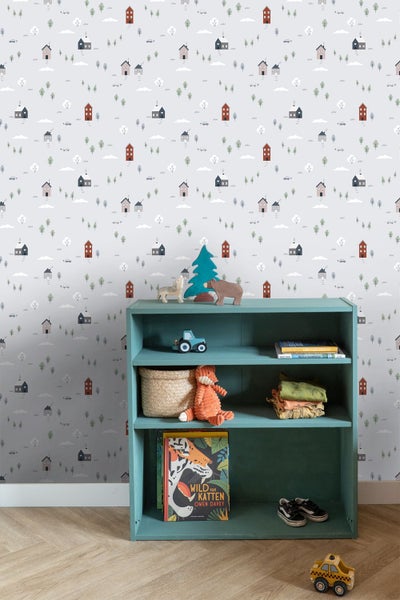 Tapete mit Häuser- und Baummotiv im Kinderzimmer, mit einem Regal voller Spielzeug und Kinderbüchern vor der gemusterten Wand.