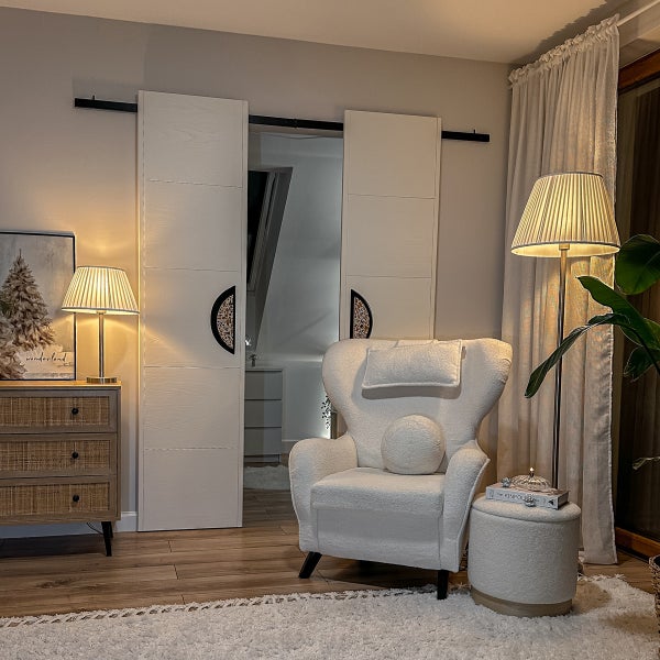 Heller Wohnraum mit weißen Schiebetüren, Bouclé-Sessel, Hocker, Stehleuchte und Holzkommode mit Rattangeflecht auf Laminatboden.