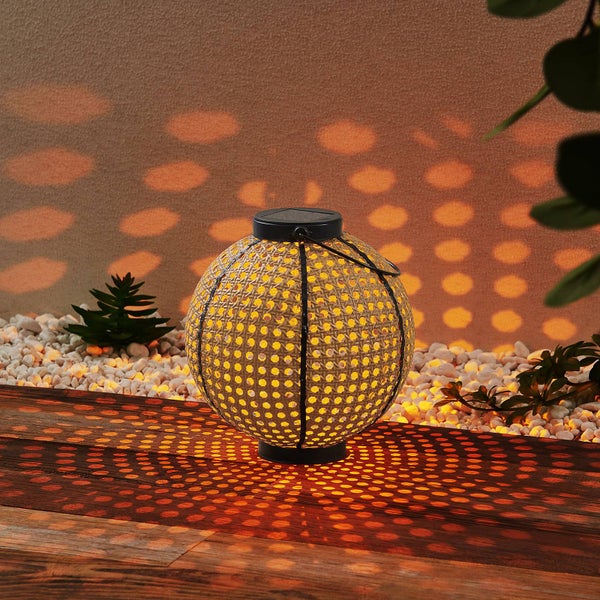 Warm leuchtende runde Solar-Laterne mit Flechtmuster auf Holzterrasse, projiziert Lichtpunkte auf Wand und weiße Steine.