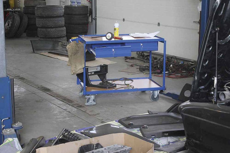 Blauer Werkstattwagen aus Metall mit zwei Ebenen und Schubladen auf Rollen in einer Garage mit Autoreifen und Ersatzteilen.