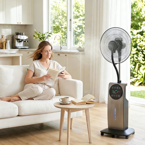 Moderner Standventilator mit Sprühnebelfunktion und Digitalanzeige im hellen Wohnzimmer neben einer Person, die auf einem weißen Sofa liest.