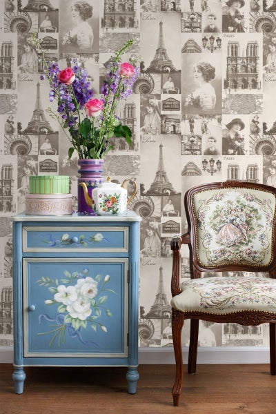Szene mit blauem Nachttisch mit Blumenmuster, Blumenstrauß in einer Vase und einem Stuhl vor einer Tapete mit Paris-Motiv