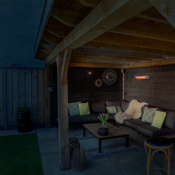 Überdachter Außenbereich mit einer Lounge-Ecke unter einem Holzpavillon. Dunkle Holzwand mit Infrarotheizstrahler, Ecksofa mit Kissen und Holztisch.